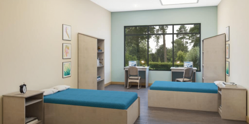 A room featuring two beds and a desk, designed for functional and durable use in behavioral health settings.